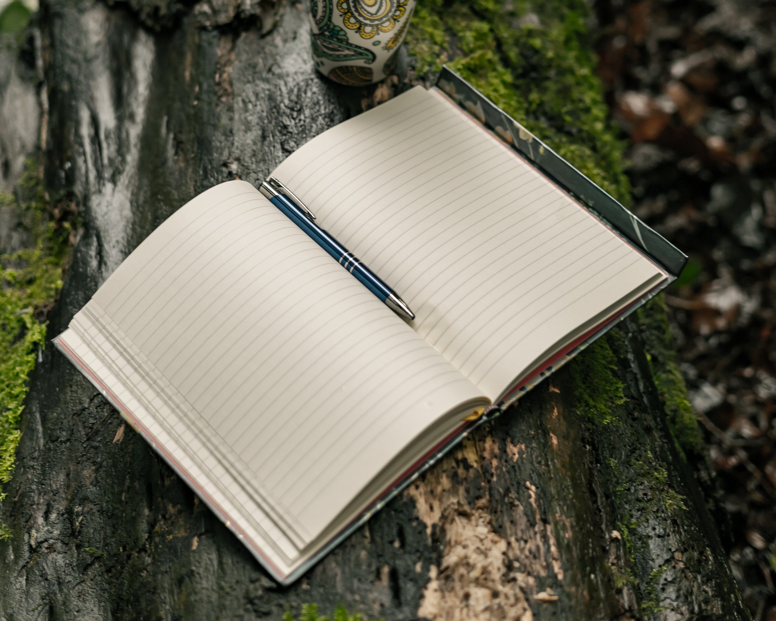 An open journal with a purple pen set between blank lined pages sits on a tree bark in a forest.