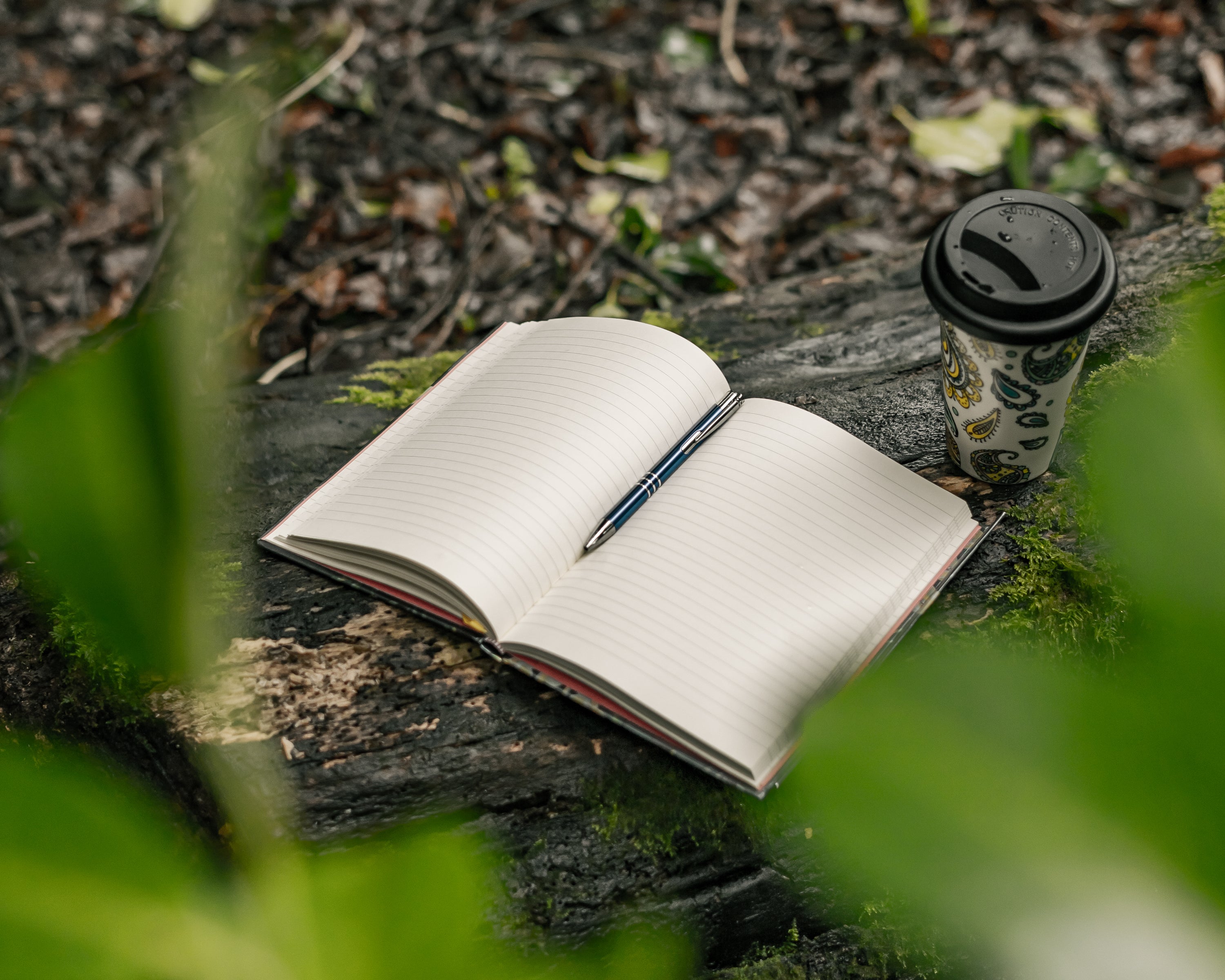 An open lined journal sits on a fallen tree, surrounded with green leaves and fresh atmosphere. A reusable glass cup is places next to it. The nature setting and the open journal looks inviting to anyone who wants to begin journaling.