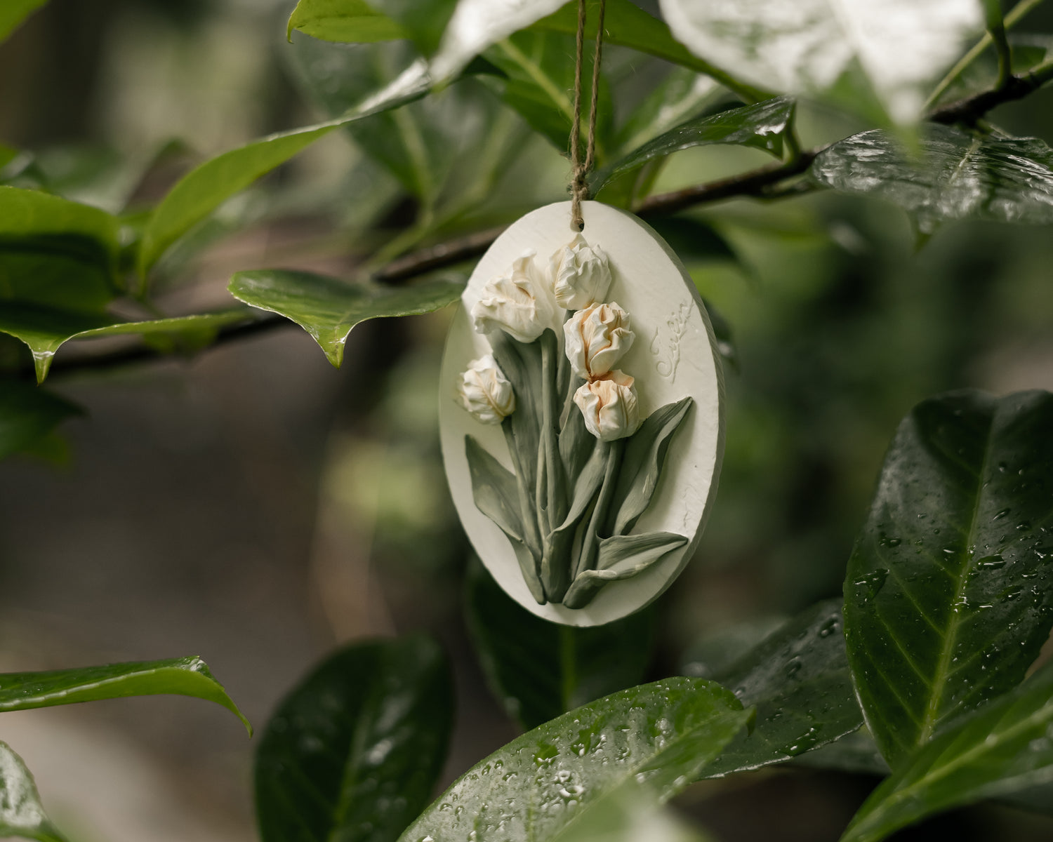 On a branch with dark green leaves with raindrops, hangs an oval handmade aroma diffuser stone with tulip moulded art.